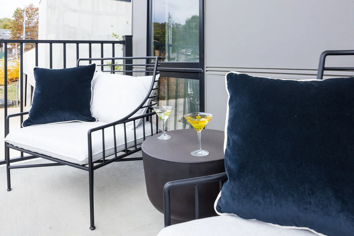 The image captures an outdoor lounge area featuring black metal chairs with plush blue pillows and cocktail glasses on the table, suggesting a comfortable space for relaxing and socializing poolside or at the BBQ area. The setting is open and inviting, perfect for unwinding or enjoying food and drinks outdoors.