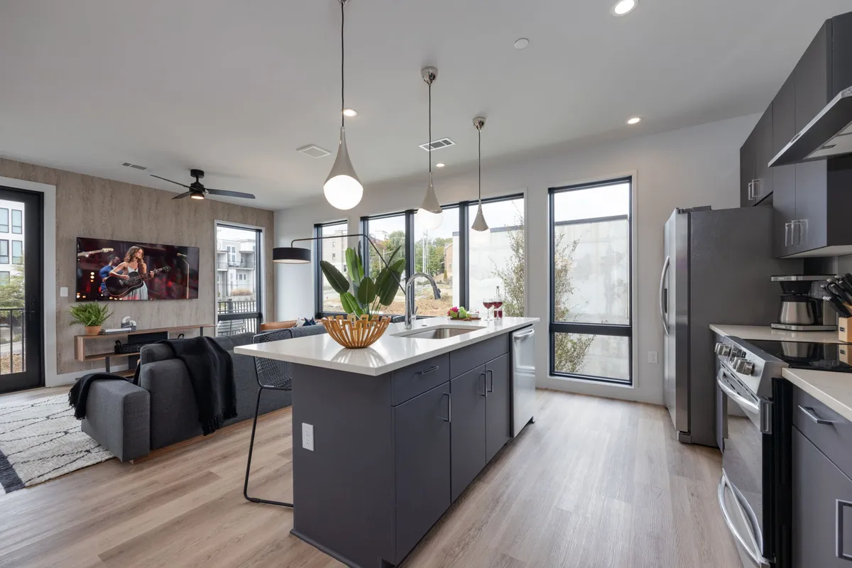 An open-plan kitchen and living space with a large island, ample natural light streaming in through expansive windows, and a connected living area. The kitchen looks fully equipped, with the flow between kitchen and dining spaces ideal for gatherings.