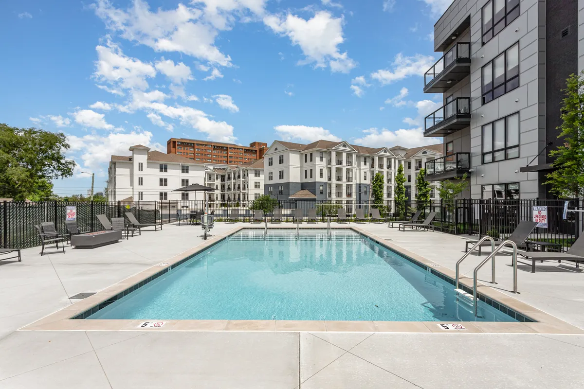 This image shows a sparkling swimming pool surrounded by modern apartment buildings, with lounge chairs and lush greenery creating a vibrant, resort-like ambiance. The pool area looks inviting for both swimming and social gatherings, with sunset lighting that highlights the year-round appeal.