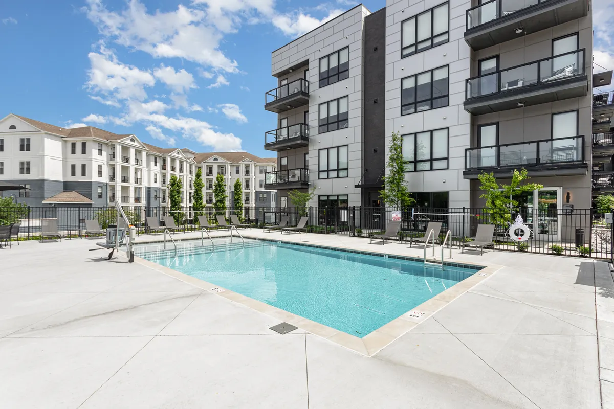 A vibrant exterior scene showing the building's swimming pool area, surrounded by green landscaping, lounge chairs, and balconies. The poolside area looks welcoming with plenty of space for groups to relax and socialize.