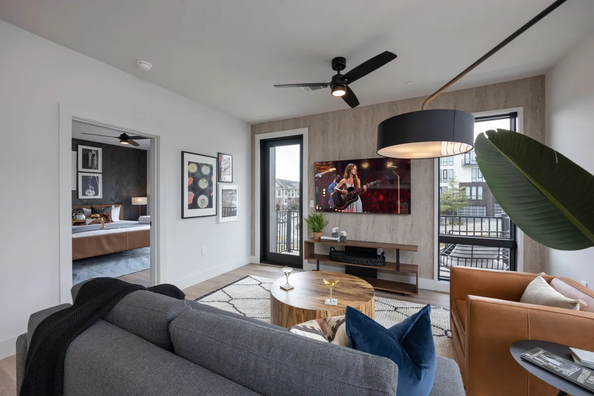 A comfortable living room featuring a sofa, coffee table, television displaying a performance, wall artwork, and large windows that flood the space with natural light. The room feels lively and is perfect for both relaxation and gatherings, with city views visible through the windows.