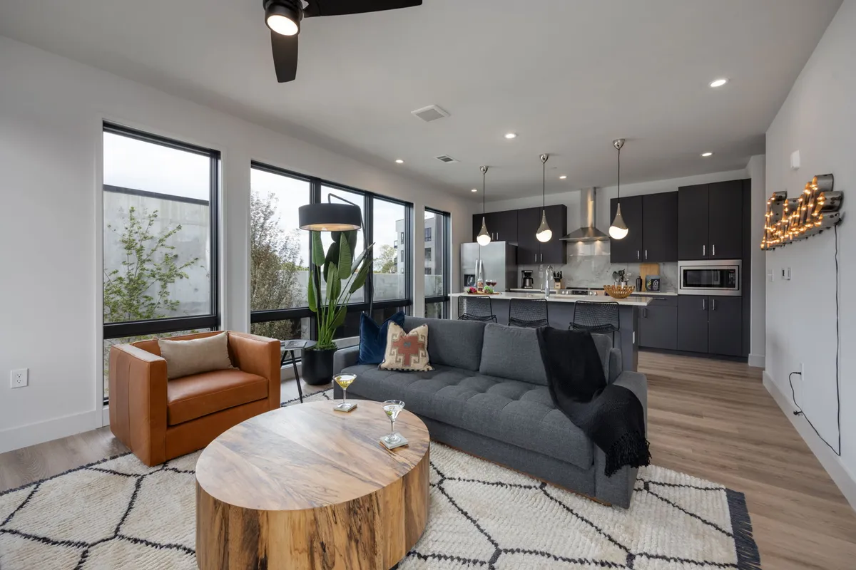 The hero image showcases a stylish, contemporary living space with a plush gray sofa, a modern wood coffee table, a bold orange armchair, and a sleek kitchen area in the background, all set within an open-concept layout. The inviting arrangement is perfect for relaxing, entertaining, or enjoying a casual meal with friends and family, reflecting the modern, cozy vibe of Nashville's Midtown.