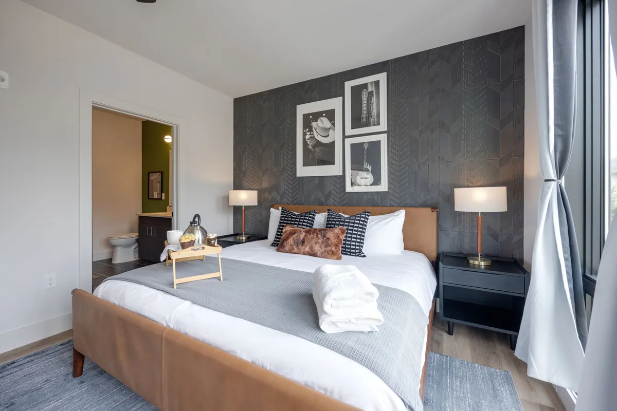 A cozy bedroom scene featuring a bed with decorative pillows, framed artwork on the wall, nightstands with lamps, and a visible bathroom door. The room is filled with natural light and has a welcoming atmosphere, perfect for restful nights or lazy mornings.