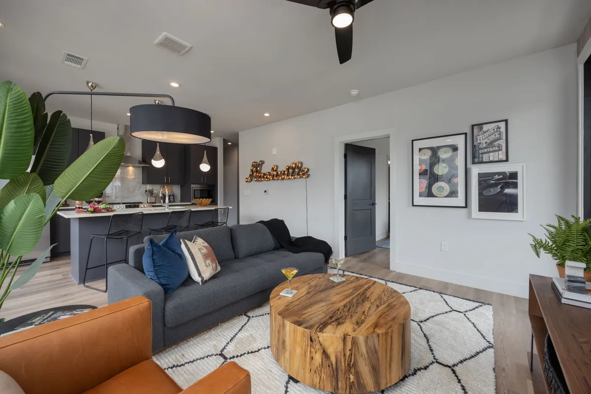 A contemporary living room with a plush sectional sofa, stylish decorative wall art, and a coffee table, with the kitchen area visible in the background. The space is open and bright, designed for relaxing, socializing, or entertaining guests.