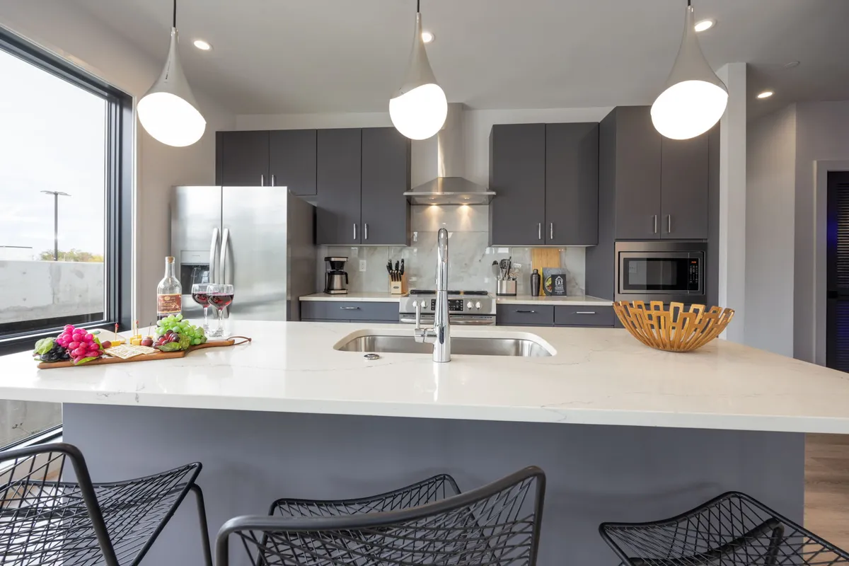 A modern kitchen island serves as the focal point, with stylish pendant lights above, sleek stainless steel appliances in the background, a bowl of fruit, and wine glasses hinting at social gatherings. The space exudes a contemporary, welcoming atmosphere, ideal for cooking, casual dining, and entertaining.