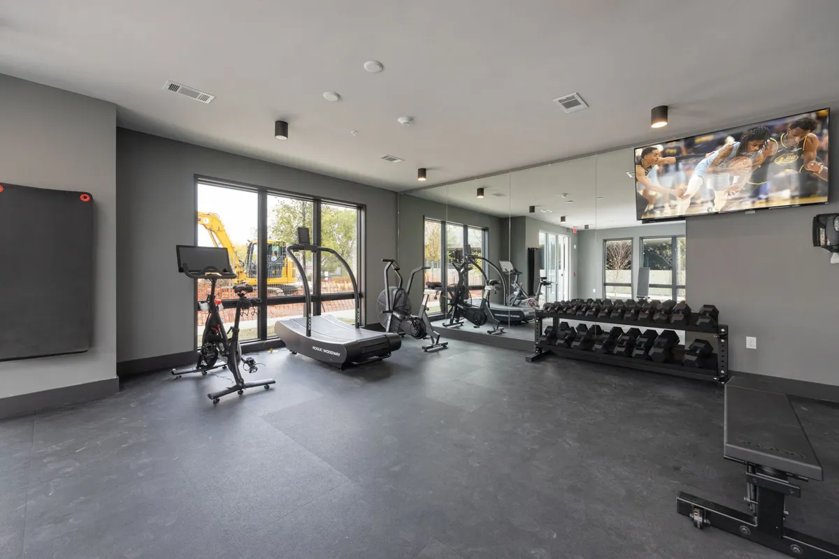 This modern fitness center features a treadmill, a dumbbell rack, large mirrors, and a TV screen, offering a well-equipped space for workouts, group fitness classes, or individual training sessions. The bright lighting and organized layout create an invigorating environment.